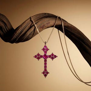May include: A cross pendant necklace featuring a series of round, vibrant pink stones set in a metallic frame. The cross is suspended from a delicate, thin gold-toned box chain, creating a structured and symmetrical jewelry piece.