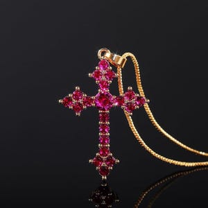 Puede incluir: Un collar dorado con un colgante en forma de cruz. La cruz está adornada con numerosas piedras preciosas rosas. La cadena también es dorada, sobre un fondo negro.