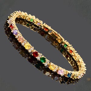 Könnte beinhalten: Ein goldfarbenes Armband mit einer Reihe farbiger Edelsteine, darunter Rot, Gelb, Grün und Blau, die mit kleinen, klaren Steinen durchsetzt sind. Das Armband liegt auf einer dunklen, reflektierenden Oberfläche.