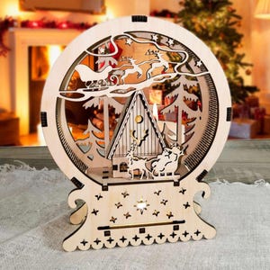 Puede incluir: Adorno navideño de madera en un marco circular, con un diseño cortado con láser del trineo de Papá Noel, renos, una casa y árboles. La base tiene recortes de estrellas, y la pieza está iluminada con una luz cálida.