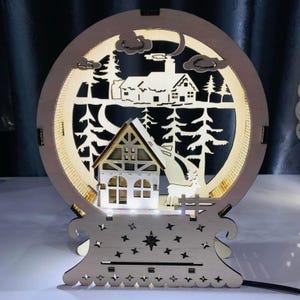 Puede incluir: Decoración navideña de madera iluminada con una escena invernal. El diseño incluye una casa, árboles, un ciervo y una luna creciente, todo dentro de un marco circular. La base tiene recortes de estrellas y forma de trineo. Alimentado por cable.