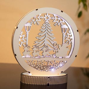 May include: A round, wooden Christmas decoration with a laser-cut design. The design features a Christmas tree, reindeer pulling a sleigh, street lamps, and snowflakes. The decoration is illuminated from within, creating a warm glow. The base is a dark brown cylinder.
