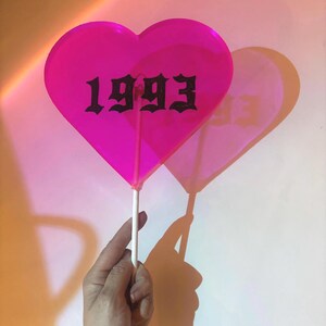 May include: A bright pink heart-shaped decoration on a white stick. The number "1993" is printed in black gothic font across the heart. The decoration casts a shadow on the white background.