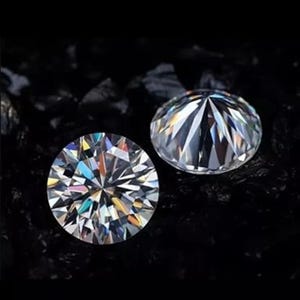Puede incluir: Dos gemas redondas y claras, con intrincadas facetas, se muestran sobre un fondo oscuro. Las gemas brillan, reflejando varios colores de luz. Las gemas son probablemente diamantes o simulaciones de diamantes.