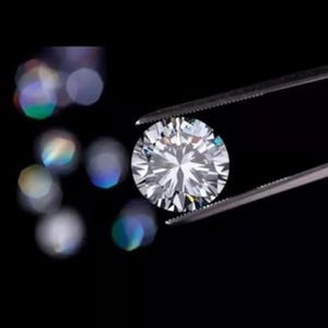 Puede incluir: Un diamante de talla brillante, sostenido por unas pinzas sobre un fondo negro. Las facetas del diamante reflejan la luz, creando un efecto brillante. El fondo est&aacute; borroso, con bokeh, atrayendo la atenci&oacute;n hacia la gema.