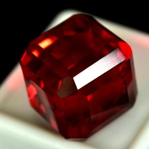 May include: A faceted, deep red gemstone with a square shape. The gem has multiple reflective surfaces, creating a brilliant display of light and color. The gemstone is set against a white surface.