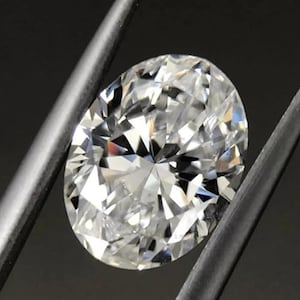 May include: A brilliant, oval-cut diamond held by a pair of tweezers. The diamond is clear and faceted, reflecting light to create a sparkling effect. The background is dark, emphasizing the diamond's brilliance.