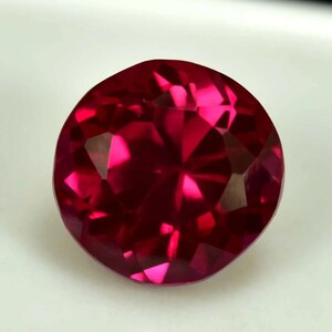 Peut inclure: Une pierre précieuse rubis ronde et facettée. La gemme est d'un rouge profond et saturé avec de multiples facettes qui réfléchissent la lumière, créant des reflets brillants. La pierre est posée sur un fond blanc.