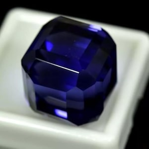 Può includere: Una gemma blu scuro sfaccettata, di forma rettangolare. La gemma è posta su uno sfondo bianco, evidenziando la sua taglio brillante e il suo colore. Le sfaccettature riflettono la luce.