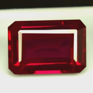 Puede incluir: Una gema rectangular facetada de color rojo intenso. La gema tiene un borde blanco y una superficie pulida que refleja la luz. El corte es esmeralda, mostrando la claridad y el color de la piedra. Adecuado para joyería.