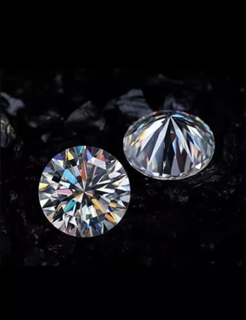 Puede incluir: Dos gemas redondas y claras, con intrincadas facetas, se muestran sobre un fondo oscuro. Las gemas brillan, reflejando varios colores de luz. Las gemas son probablemente diamantes o simulaciones de diamantes.