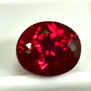 Peut inclure: Une pierre précieuse de type rubis, de forme ovale et facettée. La gemme est d'un rouge profond et saturé, avec de multiples facettes qui réfléchissent la lumière. Le rubis est posé sur un fond blanc.