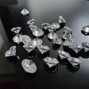 Puede incluir: Una colecci&oacute;n de cristales transparentes facetados en forma de diamante dispersos sobre una superficie negra reflectante. Los cristales, de varios tama&ntilde;os, capturan la luz y crean reflejos brillantes. El efecto general es de brillo y lujo.