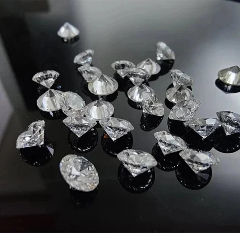 Puede incluir: Una colecci&oacute;n de cristales transparentes facetados en forma de diamante dispersos sobre una superficie negra reflectante. Los cristales, de varios tama&ntilde;os, capturan la luz y crean reflejos brillantes. El efecto general es de brillo y lujo.