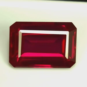 Könnte beinhalten: Ein rechteckiger, facettierter Rubin-Edelstein mit tiefroter Farbe. Der Edelstein hat ein sauberes, poliertes Aussehen mit einem weißen Rand, der seine Klarheit und Brillanz zeigt. Der Schliff ist ein Smaragdschliff mit einer symmetrischen Anordnung der Facetten.