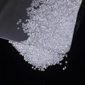 Puede incluir: Primer plano de un mont&oacute;n de peque&ntilde;as piedras preciosas claras y brillantes. Las piedras est&aacute;n dispersas sobre una superficie oscura, algunas sostenidas en un recipiente transparente y en &aacute;ngulo. Las gemas redondas reflejan la luz.