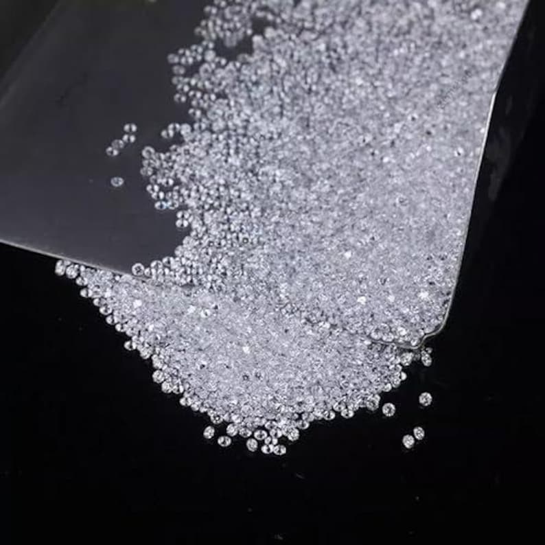 Puede incluir: Primer plano de un mont&oacute;n de peque&ntilde;as piedras preciosas claras y brillantes. Las piedras est&aacute;n dispersas sobre una superficie oscura, algunas sostenidas en un recipiente transparente y en &aacute;ngulo. Las gemas redondas reflejan la luz.