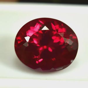 Puede incluir: Una gema de rubí tallada en forma ovalada y facetada. La gema es de un color rojo intenso y saturado con reflejos internos. Las facetas crean un juego de luces en la superficie, realzando su brillo. La gema se encuentra sobre un fondo blanco.
