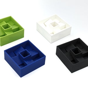 May include: Four square puzzle boxes in green, white, blue, and black. Each box has a central square and surrounding rectangular compartments. The boxes are arranged on a white surface, showcasing the geometric design and color variations.