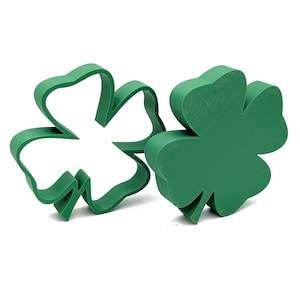 May include: Two green shamrock-shaped cookie cutters. One is a solid shamrock, and the other is the outline of a shamrock. The cookie cutters are made of a solid green material and are set against a white background.
