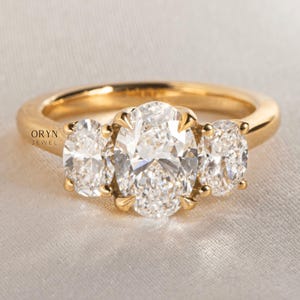 May include: A gold ring featuring three oval-cut diamonds. The center diamond is the largest, flanked by two smaller diamonds. The ring has a polished gold band and is set against a soft, neutral background. The brand name "ORYN JEWEL" is visible.