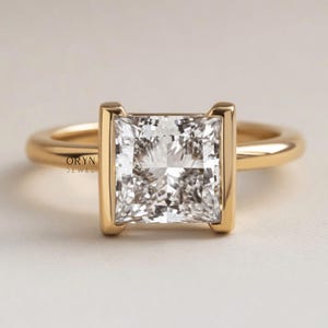 May include: A gold ring with a square, clear gemstone. The gemstone is set in a gold bezel, with a gold band. The ring is a piece of jewellery, possibly an engagement or fashion ring. The ring is marked with the text "ORYN JEWEL".