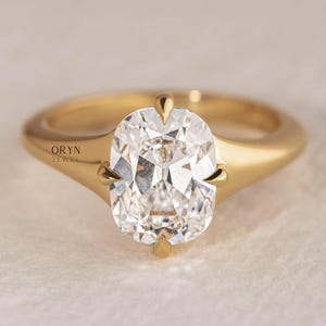May include: A gold ring with a large, oval-cut diamond. The diamond is held in place by four prongs and sparkles with many facets. The ring's band is smooth and polished, with the text "ORYN JEWEL" visible on the side.