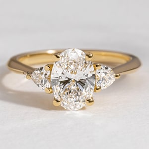May include: A gold engagement ring with a large oval-cut diamond center stone and two pear-shaped side stones. The ring has a delicate band and is set against a white background. The brand name "ORYN JEWEL" is visible.