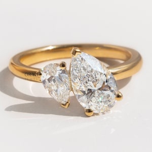 May include: A gold ring featuring two pear-shaped diamonds. The diamonds are set side-by-side, with a smaller diamond next to a larger one. The ring has a smooth, polished band and is set against a white background.