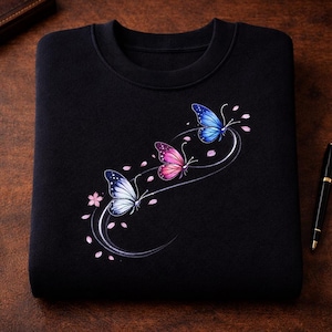 May include: A black crewneck sweatshirt featuring a graphic print of three butterflies in blue, pink, and white. The butterflies are arranged in a curved, flowing line, accompanied by small pink flower petals scattered around the design.