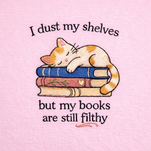 Könnte beinhalten: Gesticktes Design mit einer schlafenden orange-weißen Katze auf einem Stapel blauer, roter und beigefarbener Bücher. Der Text lautet: „Ich staube meine Regale ab, aber meine Bücher sind immer noch schmutzig“ auf rosa Hintergrund.