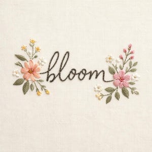 Op de afbeelding: Geborduurd kunstwerk op crèmekleurige stof met het woord 'bloom' in donkergrijs schrift. Bloemenaccenten in roze, perzik, geel en wit omringen het woord, met groene bladeren. Het ontwerp is gecentreerd.