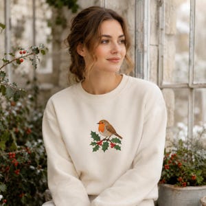 May include: A cream-colored crewneck sweatshirt featuring an embroidered graphic of a small brown bird with a bright orange chest perched on a green holly branch with red berries centered on the chest area of the garment.