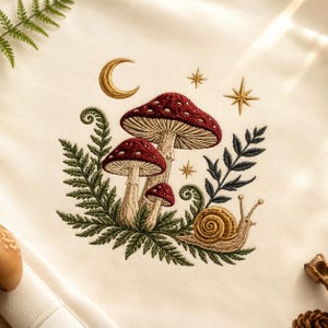 Könnte beinhalten: Ein cremefarbener Stoff mit einer Stickerei aus drei rotköpfigen Pilzen, einer Schnecke mit beigem Gehäuse, grünen Farnwedeln, einem goldenen Halbmond und zwei goldenen Sternen. Das Design ist mit detaillierten Stichen in Rot, Beige und Grün ausgeführt.