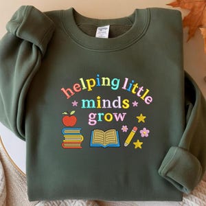 Może przedstawiać: Zielona bluza w kolorze oliwkowym z haftowanym napisem "helping little minds grow" w kolorowych literach. Projekt zawiera jabłko, książki, otwartą książkę, ołówek, kwiaty i gwiazdki. Wygodny, długi rękaw.