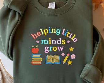 May include: Olive green sweatshirt with the embroidered phrase "helping little minds grow" in colorful letters. The design includes an apple, books, an open book, a pencil, flowers, and stars. A cozy, long-sleeved garment.