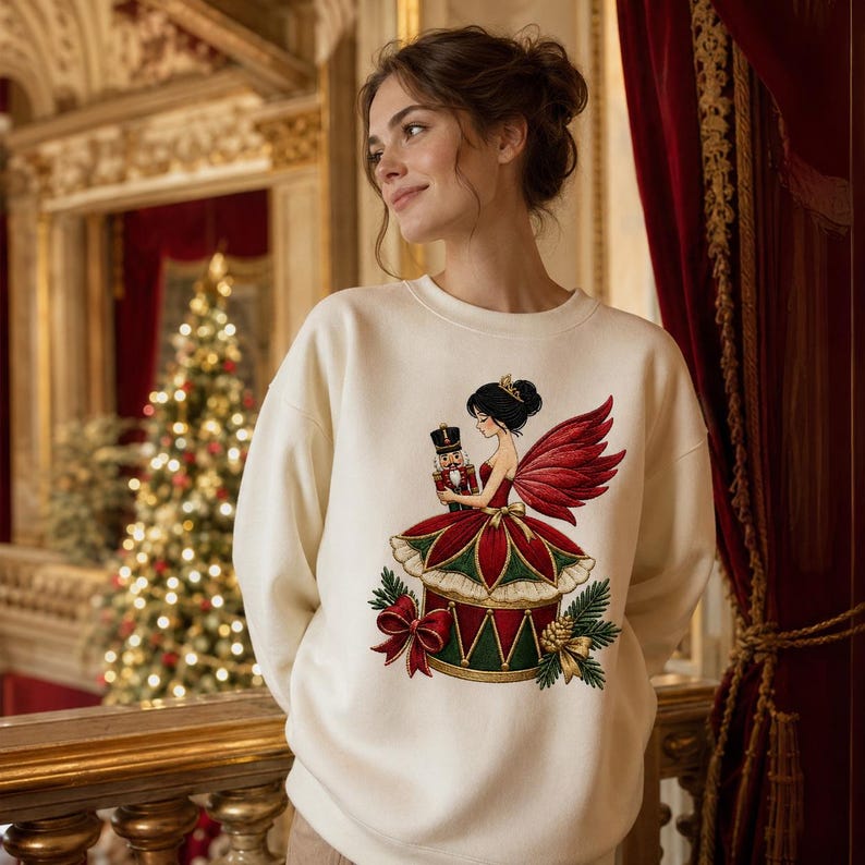 May include: A cream-colored crewneck sweatshirt featuring a graphic illustration of a fairy with dark hair and red wings, wearing a red and green dress, holding a nutcracker. The design includes pine branches, a red bow, and a gold pinecone.