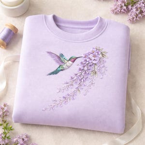 May include: A light purple crewneck sweatshirt featuring a detailed graphic of a hummingbird with iridescent green and pink feathers hovering near a cascading cluster of light purple wisteria flowers with green leaves and delicate stems.