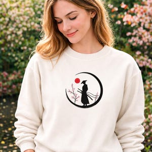 May include: Cream-colored sweatshirt featuring a black graphic of a samurai warrior within a black crescent moon, a red sun, and cherry blossoms. The samurai is depicted with two swords. The sweatshirt is made of a soft material.