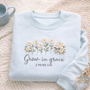 Könnte beinhalten: Hellblaues Sweatshirt mit gestickten weißen Gänseblümchen und einem Rosendesign. Der Text "Grow in grace" und "2 PETER 3:18" sind darunter aufgedruckt. Das Sweatshirt ist aus einem weichen Material.