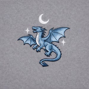 Könnte beinhalten: Gestickter Aufnäher mit einem blauen Drachen mit ausgebreiteten Flügeln, einem Mond und zwei Sternen auf grauem Hintergrund. Der Drache ist detailliert mit Schattierungen und Texturen. Das Design ist wahrscheinlich für Kleidung oder Accessoires.