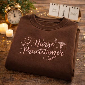 Puede incluir: Una sudadera marrón con la frase "Nurse Practitioner" en rosa, junto con símbolos médicos. La sudadera está doblada sobre una superficie de madera, con un reloj, velas y un calendario en el fondo.