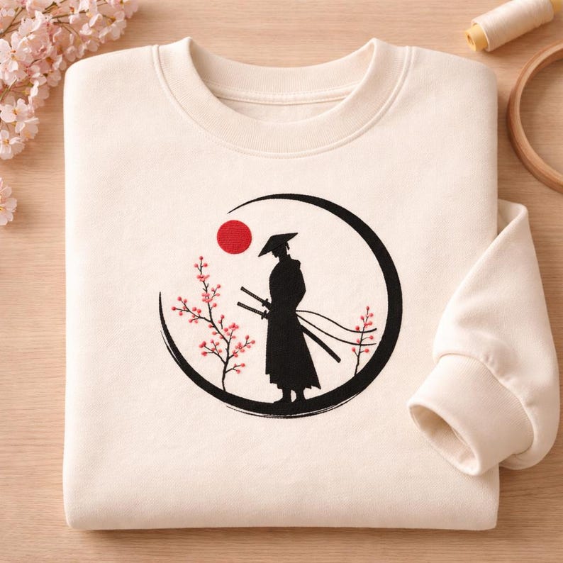 May include: Cream-colored sweatshirt featuring a black silhouette of a samurai warrior within a black circle, a red sun, and cherry blossom branches. The design includes two swords and a traditional hat. The sweatshirt has a crew neck and long sleeves.