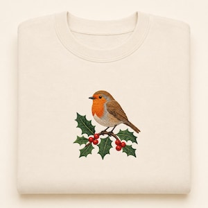 May include: A cream-colored crewneck sweatshirt featuring an embroidered illustration of a European robin perched on a holly branch. The bird has a bright orange breast and brown wings, while the holly branch displays green leaves and red berries.
