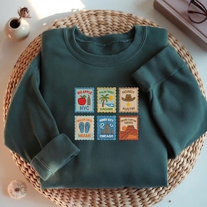 May include: Dark teal sweatshirt featuring embroidered travel-themed patches. Patches display "Big Apple NYC", "Palm Trees Sunshine", "Howdy Austin", "Beach Days Miami", "Windy City Chicago", and "Grand Canyon Views". The sweatshirt is on a woven mat.