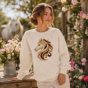 May include: A cream-coloured crewneck sweatshirt featuring an embroidered design of a horse head in profile. The horse has a flowing mane in shades of gold, brown, and cream, with intricate swirling patterns. The sweatshirt has long sleeves.