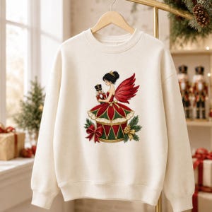 May include: A cream-colored crewneck sweatshirt featuring a central graphic of a fairy with dark hair and red wings, wearing a red and green dress, holding a small nutcracker. The design includes a red bow and pine branches at the base.