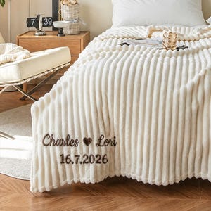 Peut inclure: Un jeté de lit de couleur crème à texture côtelée drapé sur un lit. La couverture présente une broderie marron foncé indiquant Charles et Lori avec un symbole de cœur entre les deux, suivi de la date 16.7.2026 sous les prénoms.