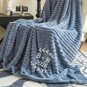 Könnte beinhalten: Eine weiche, blaue Decke, die über einem weißen Stuhl drapiert ist. Die Decke hat ein strukturiertes, geripptes Design und weiße Stickereien mit dem Text "B + H 2026". Ein gemütliches Wohnaccessoire.