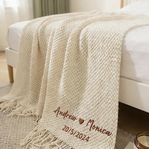 Può includere: Coperta tessuta color crema con frange, drappeggiata su un letto. La coperta presenta il testo ricamato: "Andrew ❤ Monica" e la data "20/5/2024". La coperta ha un motivo strutturato ed è realizzata in materiale morbido.